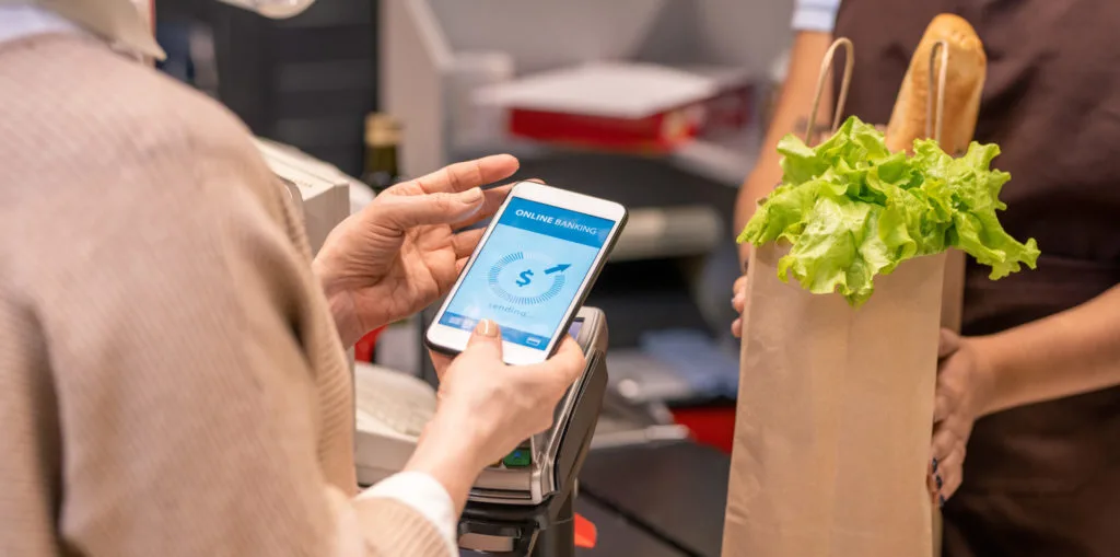 Hands of mature female buyer with smartphone over payment machine going to pay for food products in supermarket by cash register