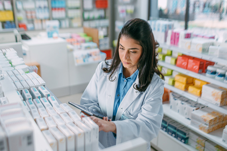 Pharmacy Drugstore: Beautiful Latina Pharmacist Uses Digital Tablet Computer, Checks Inventory of Medicine, Drugs, Vitamins, Health Care Products on a Shelf. Professional Pharmacist in Pharma Store