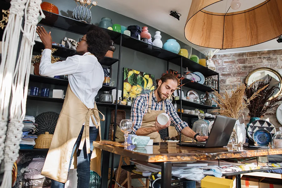 Two multicultural workers in uniform doing inventory together at decor shop. Bearded caucasian man using laptop while african woman counting goods.