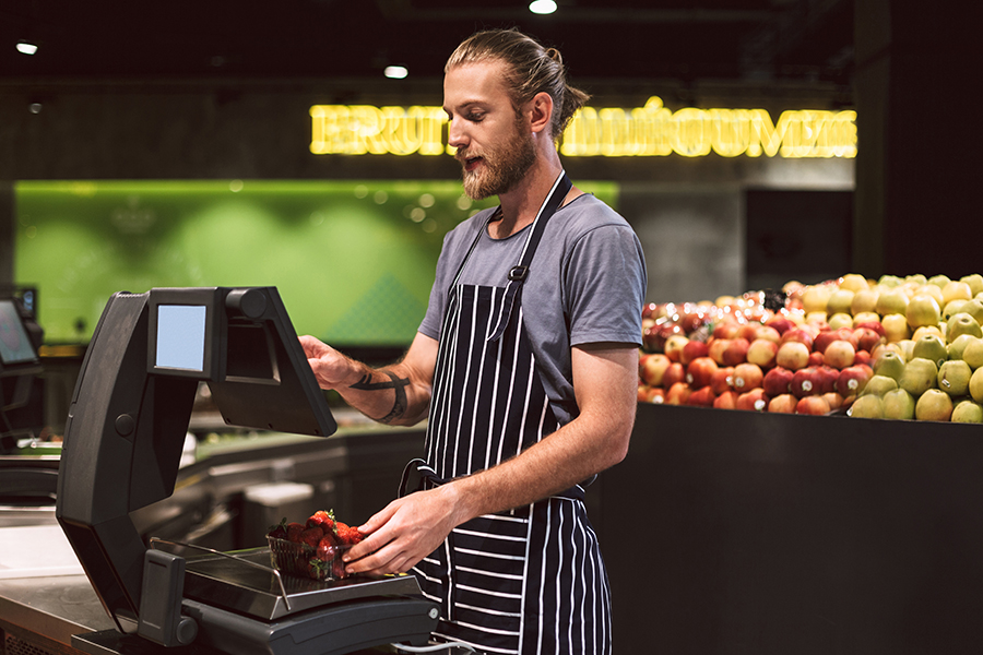Young smiling seller in apron weighing strawberries on digital scales in modern supermarket