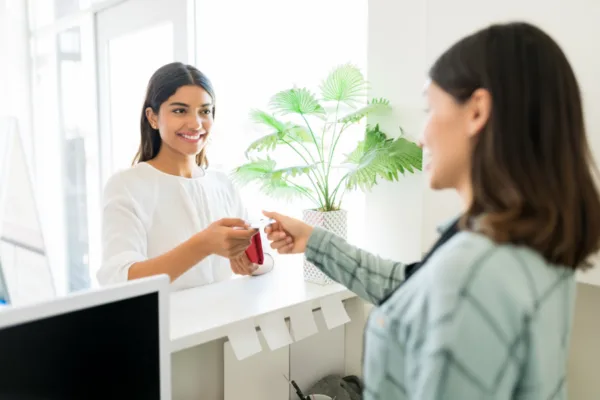 A smiling woman at a service counter hands a card to a customer standing across from her.